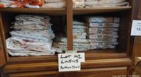 Shelf with folded and packaged vintage bed linens, labeled 'Lot 142 Linens Bottom Shelf'