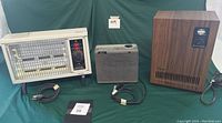 Lot of three space heaters displayed side-by-side on a table: one white radiant heater with visible heating element, one small gray fan heater, and one larger brown vintage box heater with wood pattern.