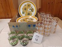 Set of 8 sunflower printed plates stacked inside a box, 8 tall drinking glasses with gold and orange patterns standing beside them, and 5 short daisy-decorated juice glasses with green and white on bases in front