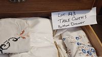 Drawer with folded tablecloths, showing various white and patterned pieces with embroidery and prints