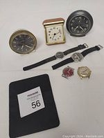 Top-down image showing three alarm clocks (brass-tone, gold-tone folding, and black round) and five wristwatches including two on straps and three cases only, all untested and in fair condition.