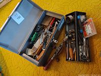 Grey metal toolbox opened showing a variety of vintage tools inside, including pliers, clamps, screwdrivers, and other hand tools with wooden and metal parts