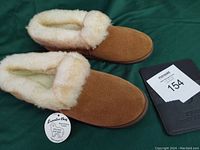 Photo shows a pair of brown suede slippers with fluffy cream wool lining inside and around the cuff. Includes a brand tag and a MaxSold lot card.