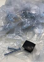 Photo of a square/rectangular cabinetry door knob in dark finish next to two silver screws on a plastic background.