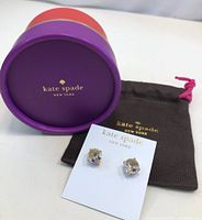 Photo of Kate Spade clear gumball stud earrings on white display card, accompanied by branded brown drawstring pouch and purple cylindrical box with Kate Spade New York logo.