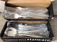 Boxes of new stainless steel forks and spoons, each piece wrapped in plastic, shown inside labeled black cardboard boxes.