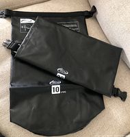 Two black waterproof bags labeled 10 liters and 20 liters respectively laid on a couch, both with roll-top closure and clip fasteners.