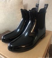 Side angled view of new black ankle rain boots on cardboard box, showing glossy finish and elastic side panel.