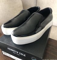 Side angle view of black patent slip-on shoes with thick white platform soles on Kenneth Cole branded box.