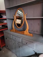 Oval wooden framed antique dresser mirror placed on a grey shelf against a wall. Shows full front and side angle of the mirror's wooden frame and mounting supports.
