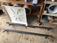 Photo showing white plastic washing machine tray with a built-in drainage hole and two black metal bed frame rails with caster wheels attached.