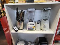 Photo of two kitchen blenders, knife block set with several knives, manual can opener, and metal utensil inside a white shelf.