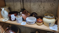 Shelf with various crocks, pitchers, and bowls including a white pitcher, several brown stoneware crocks, and ceramic bowls.
