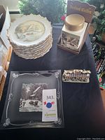 Stack of eight Currier and Ives collector plates with winter scenes and gold trim, instant coffee container, glass plate, and small decorative item on black tablecloth.