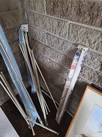 Assorted white and silver curtain rods and tension rods leaning against a brick wall, some rods in blue packaging and one in original printed packaging, all mostly new.