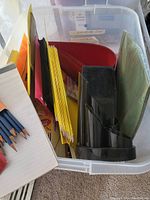 Clear plastic bin containing colored hanging file folders, black pen/pencil holder, and other miscellaneous desk supplies.