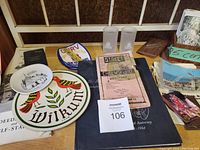 Overview of assorted ephemera on table including plates, glasses, patch, printed paper items such as postcards, labels, and folded maps.