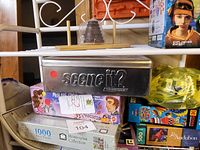 Photo showing stacked games on white metal shelving, including Scene It?, 1000 piece puzzle, and board games with cartoon characters