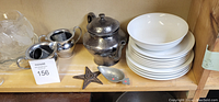 Shelf containing a pewter teapot, matching creamer and sugar bowl, stacked white ceramic bowls and plates, clear glass plates and dessert dishes, a metal star-shaped trivet, and a metal scoop.