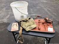 All items on display: white plastic bucket, canvas tool bag, leather hammer holster, brown tool belt piece, unopened box of nails in background