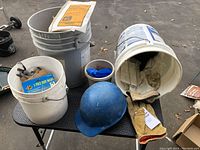 Photo of buckets containing gloves, dust masks, a blue hardhat, and safety gear items arranged on a table.