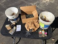 Full lot overview showing two buckets, one box and multiple small bags of nails and screws on a metal surface.