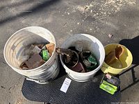 Two white plastic buckets and a small yellow bucket placed on ground, filled with assorted screws, nails, bolts, and packaging materials