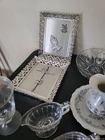 Photo showing silver-tone framed plaque with praying hands and angel, decorative cross with number 25, and parts of glassware items on a black table.