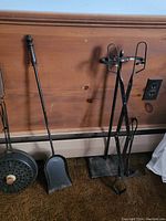 Vintage fireplace tools against a wood panel wall, showing black metal shovel and tripod stand with attached warmer pot.