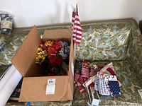 Box containing various faux flowers in red, yellow, purple, and green. Several small American flags on sticks inside and next to the box on a green cushion with floral pattern.
