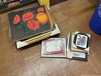 Stack of paintings with prominent oil painting of red flowers and orange fruit on top, and assorted prints and framed pictures to the side, all placed on a wooden floor.
