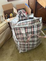 Photo of a large tartan plaid tote bag with handles filled with assorted smaller bags inside, placed on carpeted floor near furniture and cardboard boxes.