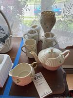 Photo showing the Belleek tea set including teapot, creamer, four teacups, and vase partially wrapped in bubble wrap on a table near a window with lace curtains.