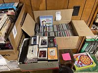 Photo showing open cardboard boxes containing a mix of CDs and cassette tapes, some in cases and some loose. Visible cassette cases have handwritten labels.