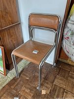 Two metal frame stacking chairs with wooden seats and backs photographed indoors against a white wall and wood parquet floor.