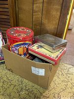 Box of assorted advertising food tins with red floral round tin, rectangular tins, and other varied shapes.