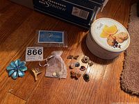 Photo showing a decorative tin containing various pins and earrings, a blue flower brooch, a yellow leaf pin, several small pins, and a small mesh bag with jewelry items inside.