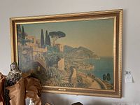Framed print featuring a Mediterranean coastal village scene with warm tones in an ornate gold frame.