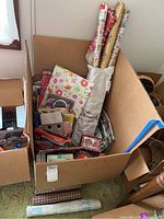 Box containing multiple rolls of gift wrapping paper, several gift bags, and gift boxes visible inside a cardboard box.