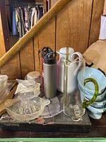 Photo of assorted kitchen and drinkware items on table including glass juicer, crystal dish, drink bottles, coffee carafe, and bowls