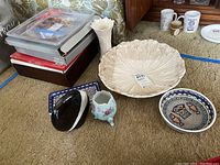 Wide view of large decorative cream-colored bowl with leaf pattern, two vases (white wavy rim, floral three-legged), black swan figure, two mosaic ceramic bowls, and boxed platters stacked in plastic containers