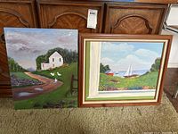 Both oil paintings displayed side by side on carpet, one framed with a coastal scene and one unframed showing a rural house.