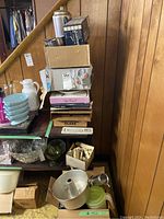 Photo showing stacked boxes containing various kitchen items including boxed flatware, a coffee percolator, plastic containers, and ceramic bowls beside a wooden wall.