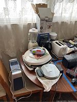Photo showing six small kitchen appliances on a table by window, including blender, waffle maker, toaster, kettles and air purifier heater