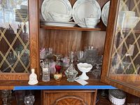 Photo of a wooden cabinet with shelves holding white porcelain plates and teacups, assorted clear glass stemware, glass bowls, small bells, and decorative items.