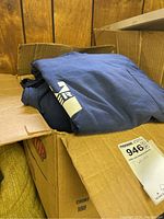 Photo of box filled with folded dark blue T-shirts, some showing partial white screen print graphics such as 'HYSTER' and large 'H'.