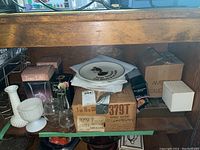 Various glassware and boxed vases placed on a wooden shelf under a table, including hobnail glass vases, milk glass vase, and several boxes.