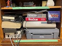 Wooden shelf holding various vintage computer electronics including Commodore 1541 drive, MPS 802 printer, VirusScan software box, SAT prep book, and arcade joystick box.