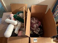 Two open cardboard boxes containing multiple skeins of yarn in various colors and green plastic round looms inside the boxes.