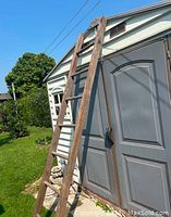 Full view of the antique tall wooden ladder leaning against a building outdoors, showing entire length and structure.